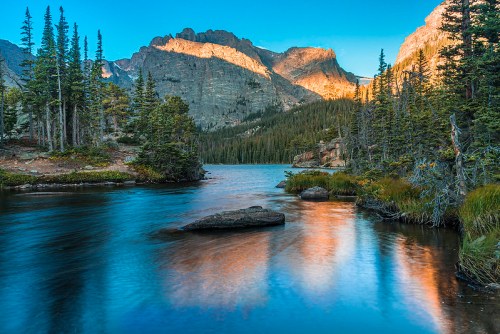 A serene lake surrounded by tall trees and mountains, reflecting the warm glow of sunset on the water's surface.