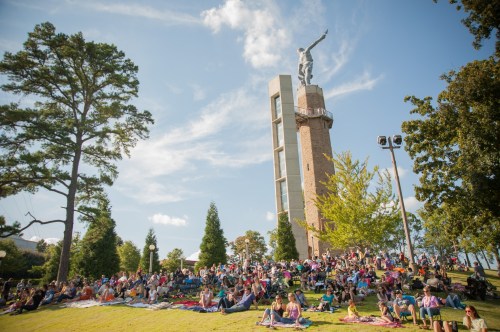 A large crowd gathers on a grassy hill near a tall monument under a blue sky with scattered clouds.