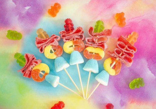 Colorful candy lollipops with gummy bears and fruit-shaped gummies on a vibrant, tie-dye background.