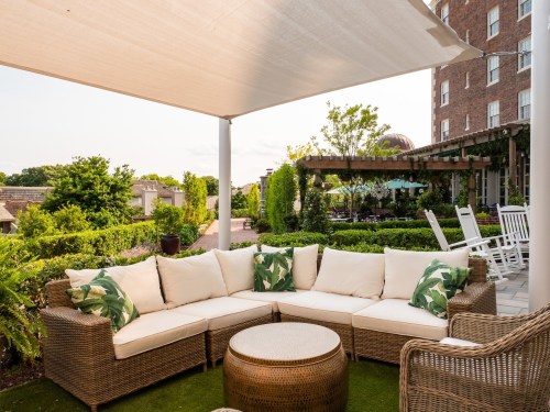 A cozy outdoor seating area with a sectional sofa, green plants, and a round table under a shaded canopy.