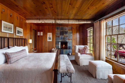 Cozy wooden bedroom with a large bed, armchair, and fireplace, featuring warm tones and natural light from windows.
