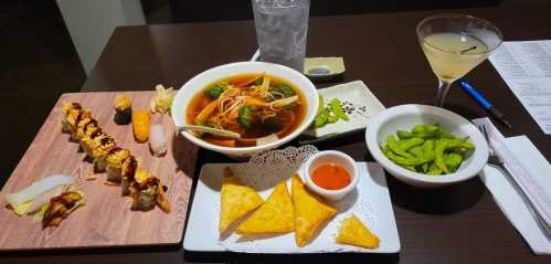 A table set with sushi, a bowl of soup, edamame, crispy snacks, and a cocktail, with a glass of water in the background.