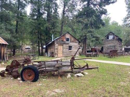 A rustic scene featuring an old plow in front of a wooden cabin, surrounded by trees and vintage farm equipment.