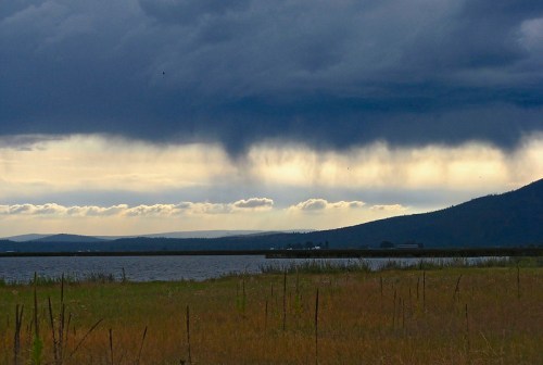 Dark clouds loom over a tranquil lake, with distant hills and a hint of sunlight breaking through the stormy sky.