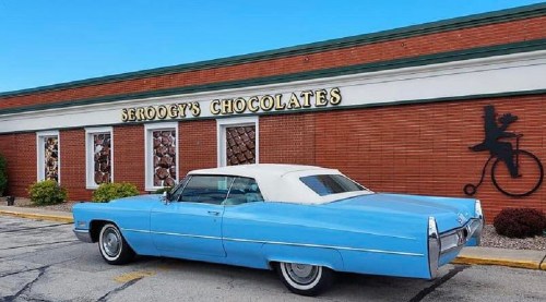 A vintage blue Cadillac parked outside Seroggy's Chocolates, featuring a brick building and a whimsical chocolate-themed mural.