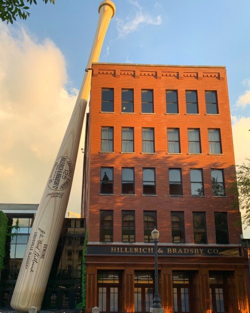 A large baseball bat leans against a brick building with large windows, labeled "Hillerich & Bradsby Co." under a blue sky.