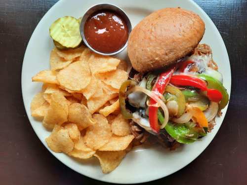 A plate with a sandwich topped with peppers and onions, served with chips, a pickle, and a small cup of sauce.