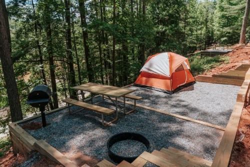 A campsite in a wooded area featuring an orange tent, picnic table, grill, and fire pit on gravel ground.