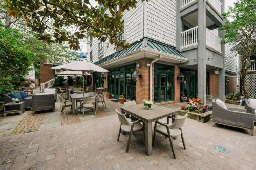 A cozy outdoor patio with tables, chairs, and greenery, surrounded by a building and colorful flowers.