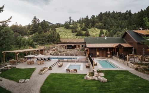 A scenic view of a lodge with pools, surrounded by trees and mountains, featuring outdoor seating areas.