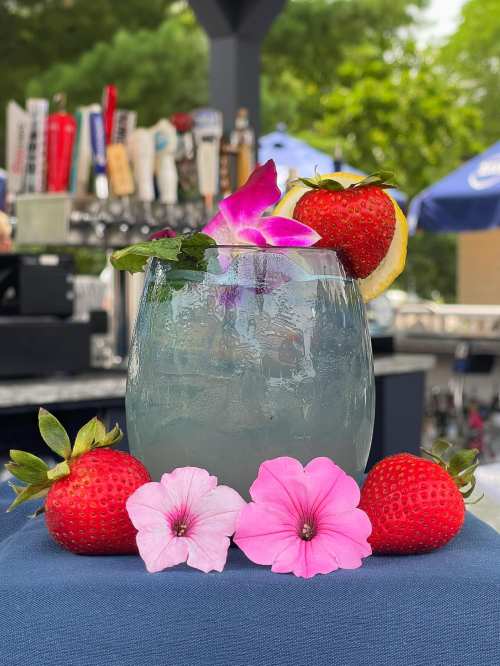 A refreshing cocktail garnished with strawberries, lemon, and flowers, set against a blurred bar background.