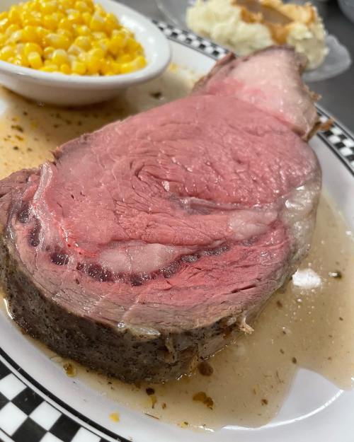 A juicy slice of prime rib served with a side of corn and mashed potatoes on a white plate.