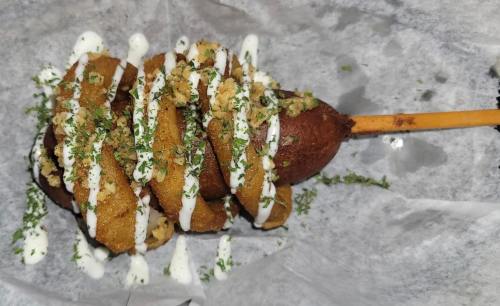 A close-up of a corn dog topped with drizzled sauce, chopped herbs, and crushed toppings on a paper-lined plate.