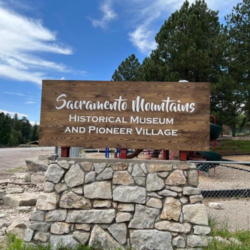 Sign for Sacramento Mountains Historical Museum and Pioneer Village, set against a backdrop of trees and playground equipment.