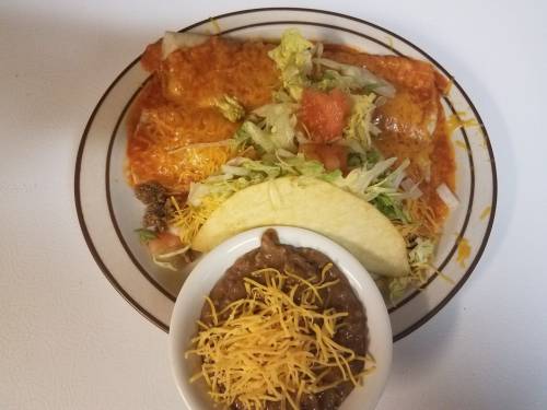 A plate with enchiladas topped with cheese, lettuce, tomatoes, a taco, and a bowl of refried beans with cheese.