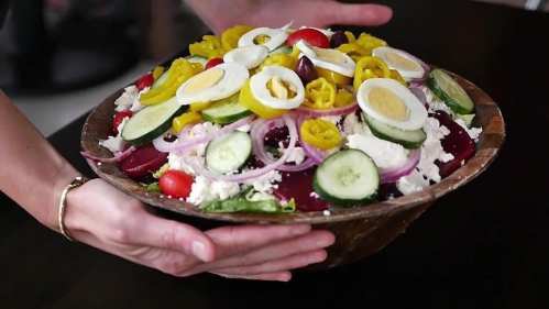 A person holds a large wooden bowl filled with a colorful salad featuring greens, tomatoes, cucumbers, and sliced eggs.