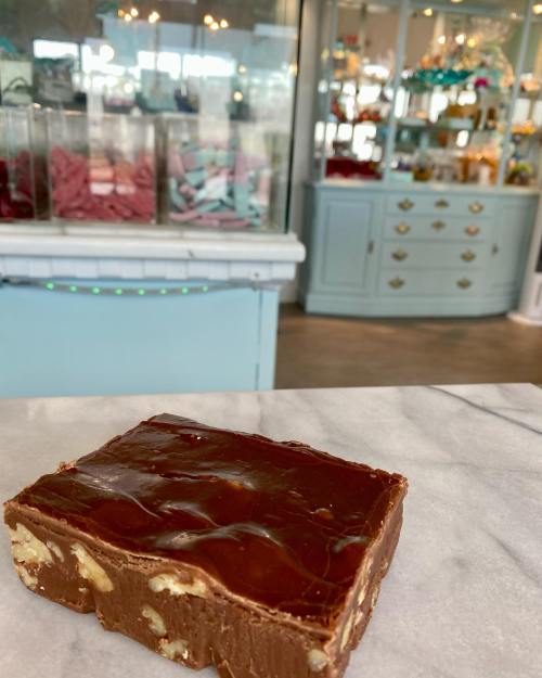 A piece of chocolate fudge with nuts sits on a marble counter, with a colorful candy display in the background.