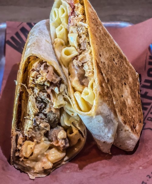 A close-up of a grilled burrito filled with macaroni, cheese, and shredded meat, resting on a red paper surface.