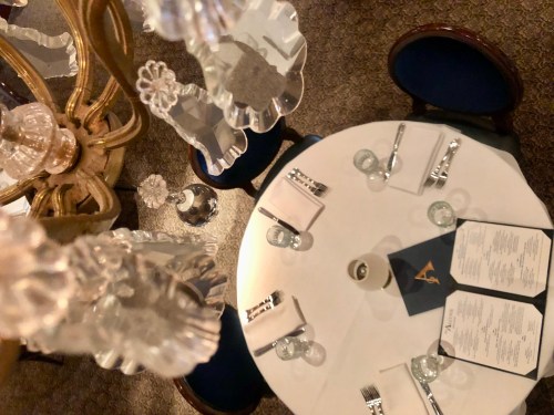 Aerial view of a dining table set with menus, glassware, and silverware, beneath an ornate chandelier.