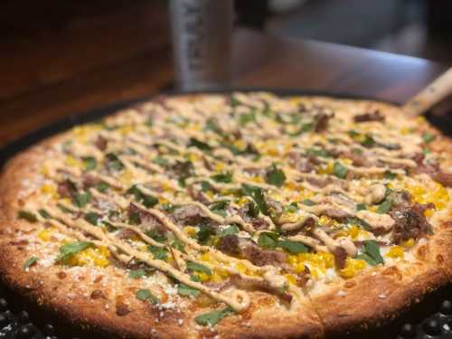 A close-up of a pizza topped with meat, corn, herbs, and drizzled with sauce on a wooden table.