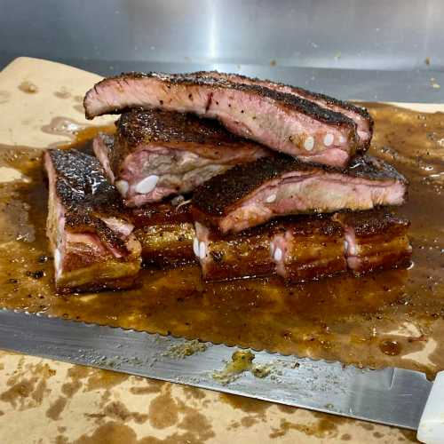 A stack of smoked, juicy ribs with a crispy crust, resting on a metal surface with a sheen of sauce.