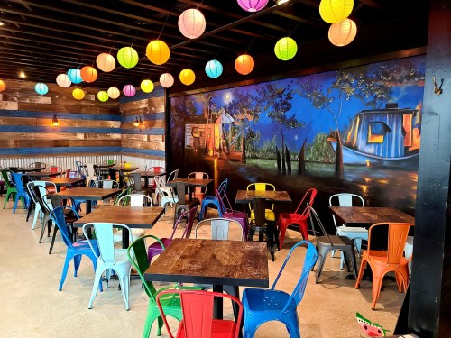 Colorful chairs and tables in a vibrant restaurant with lanterns and a mural of a boat on a scenic backdrop.