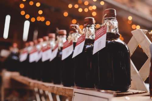 A row of glass bottles filled with dark liquid, each with a tag, displayed on a wooden shelf with warm lights in the background.