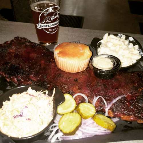 A plate of ribs with coleslaw, pickles, onions, macaroni, a muffin, and a glass of beer.