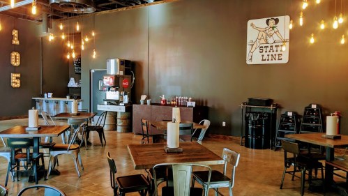 Interior of a restaurant featuring wooden tables, metal chairs, and a vintage sign, with warm lighting and a soda machine.