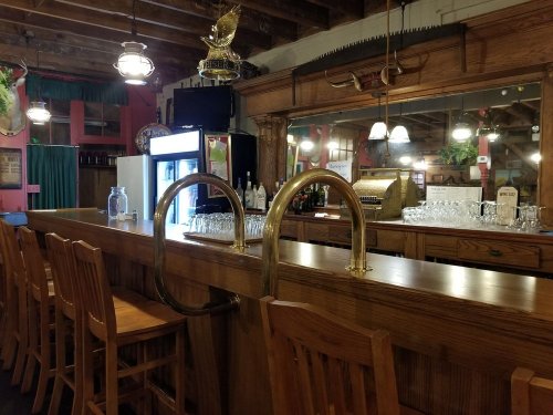 A rustic bar with wooden stools, a polished counter, and vintage decor, featuring a mirror and glassware in the background.