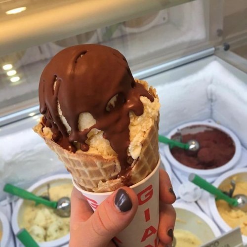 A hand holding a waffle cone with two scoops of ice cream, topped with chocolate sauce, in front of ice cream tubs.