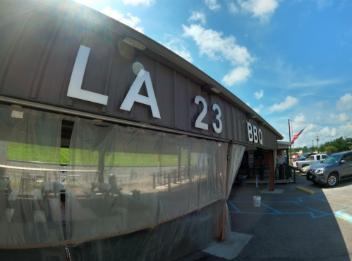 Exterior of LA 23 BBQ restaurant with large letters, blue sky, and parked cars in the foreground.