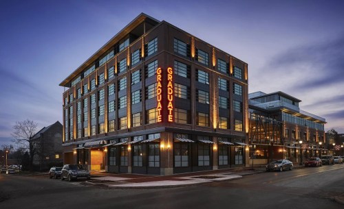 A modern hotel building with "GRADUATE" signage, illuminated at dusk, surrounded by parked cars and city streets.