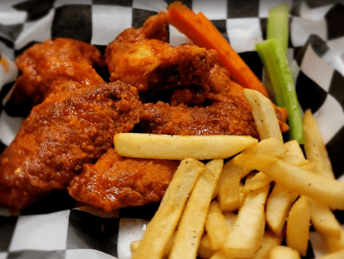 A basket of crispy chicken wings with French fries and carrot and celery sticks, served on a checkered paper.