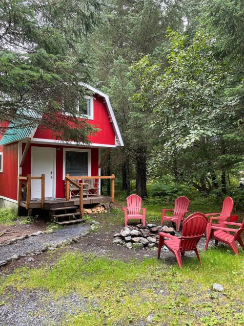 A red cabin surrounded by trees, with a small porch and a fire pit featuring red chairs in front.