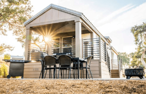 A modern tiny house with a porch, outdoor seating, and a grill, surrounded by trees and sunlight.