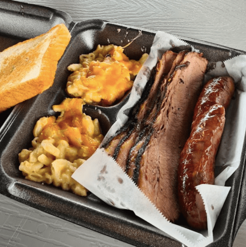 A takeout container with brisket, sausage, macaroni and cheese, and a slice of toast.