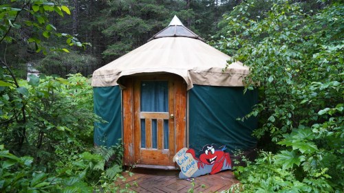 A cozy yurt surrounded by greenery, featuring a wooden door and a colorful sign at the entrance.