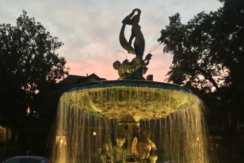 A decorative fountain with a statue of a couple dancing, surrounded by water and trees at sunset.