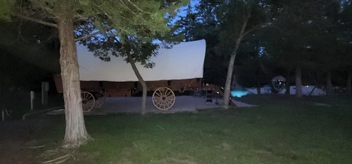 A covered wagon parked under trees at dusk, with a faint glow from nearby lights in the background.