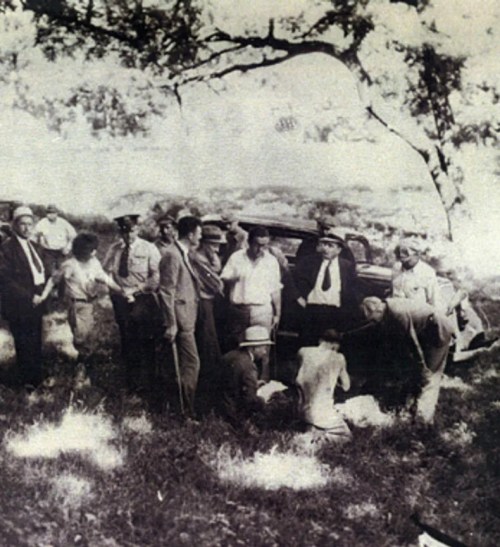 A historical black-and-white photo of a group of people gathered outdoors near a car, some observing a seated figure.