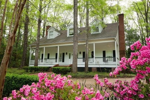 A charming white house with a porch, surrounded by tall trees and blooming pink flowers in a serene setting.