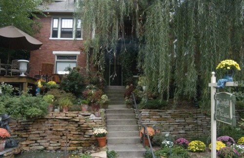 A charming brick house surrounded by lush greenery and colorful flowers, with a stone pathway leading to the entrance.