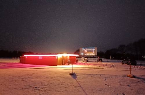 A drive-in movie theater at night, with a glowing concession stand and a large screen displaying a film, surrounded by snow.