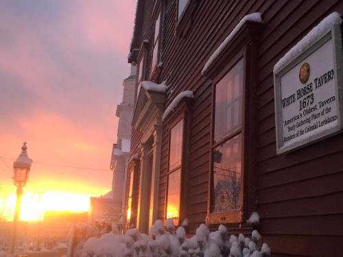 A historic tavern with a sign, surrounded by snow, as a colorful sunset lights up the sky.