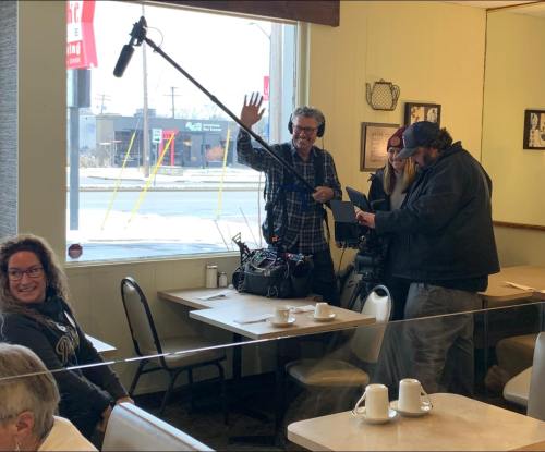 A film crew sets up in a diner, with a microphone boom and cameras, while a woman smiles in the foreground.