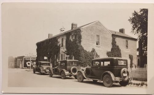 Black and white photo of vintage cars parked outside a brick building covered in ivy, with a tavern sign in the background.