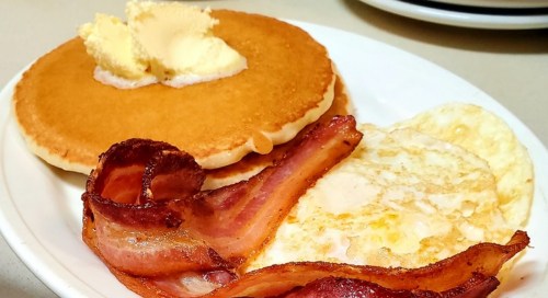 A plate with a fluffy pancake topped with butter, alongside crispy bacon and a fried egg.