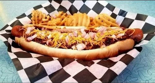 A loaded hot dog topped with chili, cheese, and onions, served with waffle fries in a checkered basket.