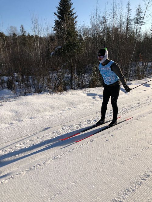 A skier in a blue bib glides over a snowy trail, surrounded by trees under a clear blue sky.